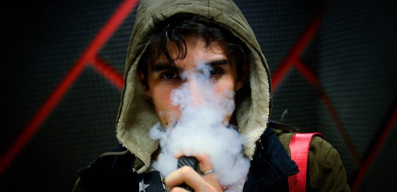 Person using a disposable vape with a large vapor cloud while wearing a hooded jacket indoors.