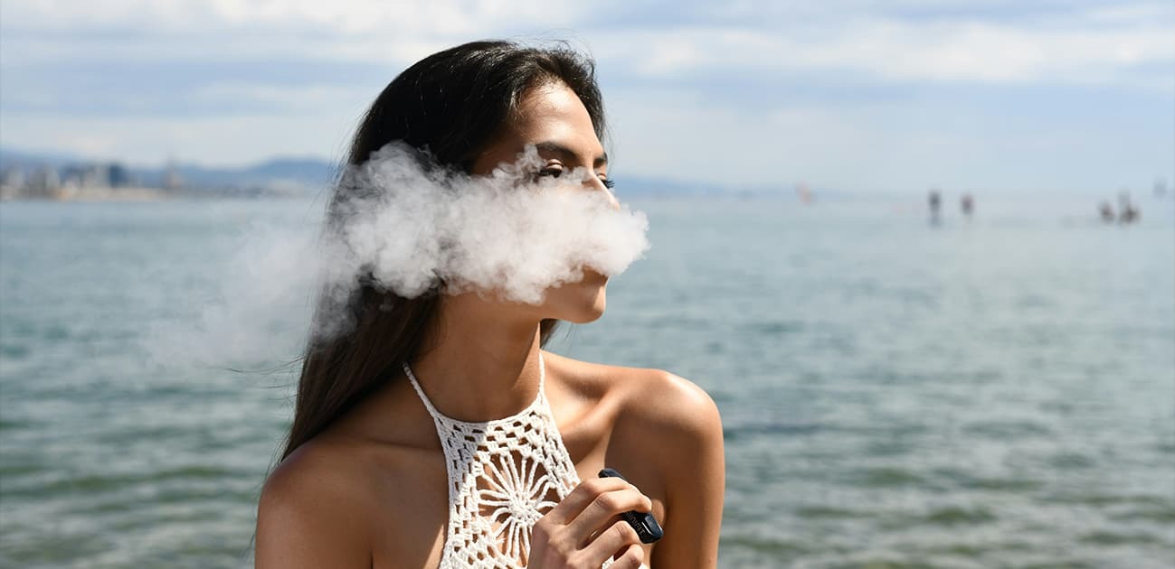 Woman vaping outdoors near the ocean with a visible vapor cloud and disposable vape device in hand.